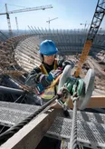 A person wearing safety equipment uses an angle grinder on a steel beam at a stadium construction site.