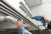 A person drills into a ceiling-mounted metal channel in an industrial setting.