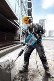 A person wearing safety equipment operates a breaker to demolish concrete at a construction site.