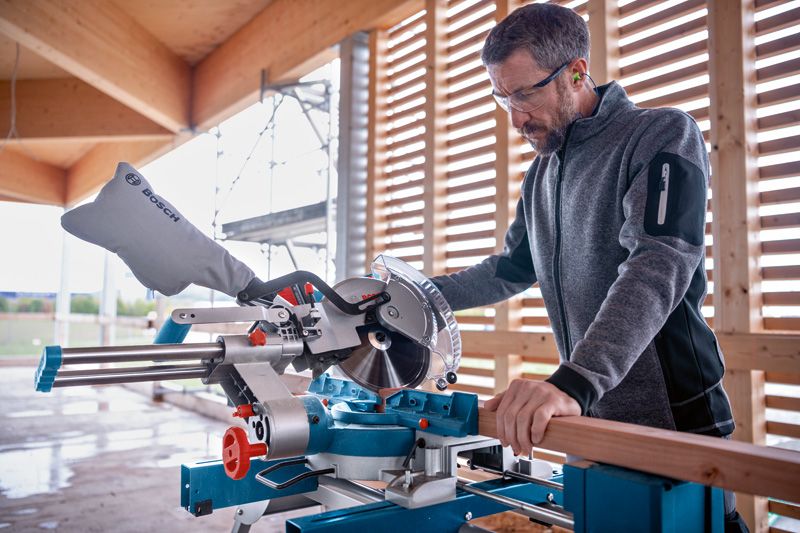 A person wearing safety equipment uses a cordless mitre saw to cut wood.