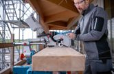 A person wearing safety equipment operates a cordless mitre saw to cut a wooden beam.