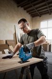 A person wearing safety equipment uses a jigsaw to cut wood on a workbench.