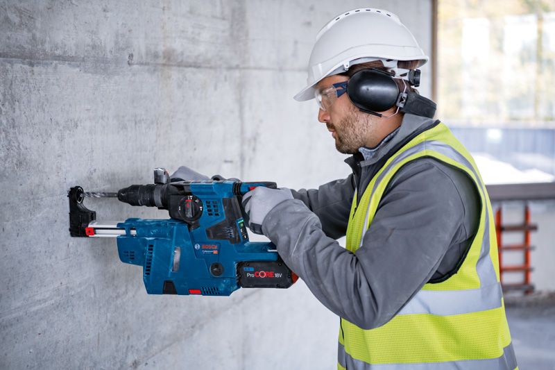 A person wearing safety equipment drills into a concrete wall with a cordless rotary hammer.