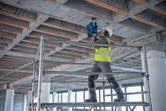 A person wearing safety equipment drills into a concrete ceiling at a construction site.
