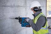 A person wearing safety equipment drills into a concrete wall with a cordless rotary hammer.