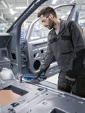 Person wearing safety equipment uses a power tool on a car frame in an industrial setting.