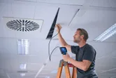 A person on a ladder inspects a ceiling cavity with an inspection camera.