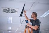 A person on a ladder inspects a ceiling panel with an inspection camera.