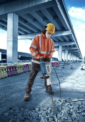 A person wearing safety equipment uses a demolition hammer to break concrete on a road.