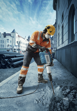 A person wearing safety equipment uses a demolition hammer to break concrete on a city street.