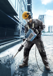 A person wearing safety equipment breaks concrete with a demolition tool at a construction site.