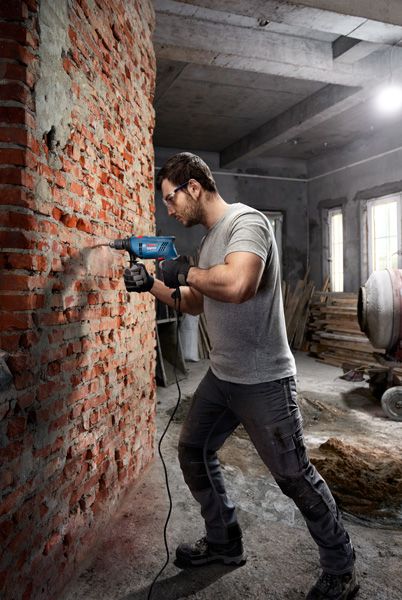 A person wearing safety equipment drills into a brick wall with an impact drill.