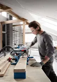 A person wearing safety equipment cuts a wooden board with a circular saw in a workshop.