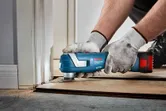 A person wearing safety equipment uses a cordless multi-cutter to trim flooring near a door.