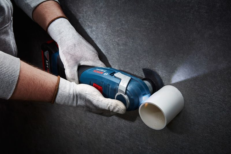 A person wearing safety equipment cuts a white plastic pipe with a cordless multi-cutter.