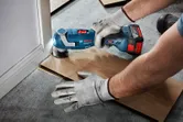 Person wearing safety equipment uses a cordless multi-cutter to trim flooring near a wall.
