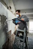 A person wearing safety equipment removes tiles from a wall using a rotary hammer.