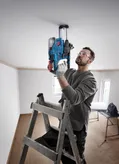 A person wearing safety equipment drills into a ceiling while standing on a ladder.