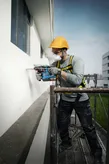 A person wearing safety equipment drills a hole into a building exterior while on scaffolding.