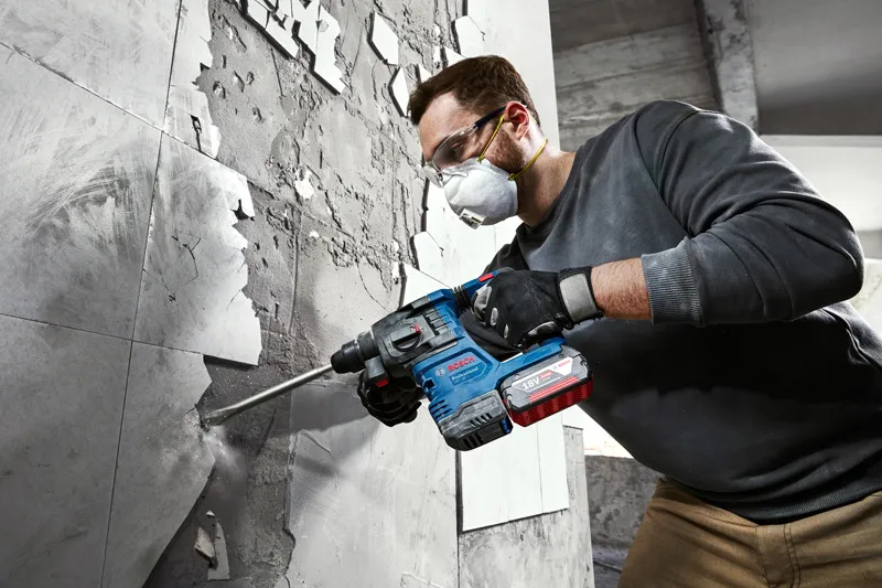 A person wearing safety equipment uses a rotary hammer to remove tiles from a wall.