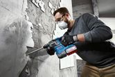 A person wearing safety equipment uses a rotary hammer to remove tiles from a wall.