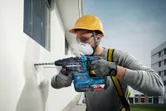 A person wearing safety equipment drills into an exterior wall with a power tool.