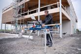 A person wearing safety equipment cuts wood with a cordless mitre saw at a construction site.