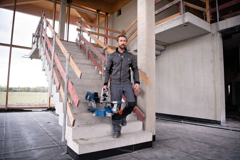 A person carrying a cordless mitre saw walks down stairs at a construction site.