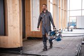 A person wearing safety equipment carries a cordless mitre saw in a construction area.