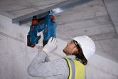 A person wearing safety equipment secures a metal channel to a concrete ceiling with a cordless concrete nailer.