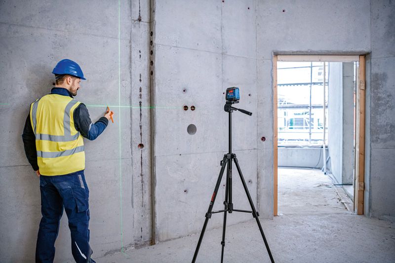A person wearing safety equipment marks a concrete wall using a laser leveling tool on a tripod.
