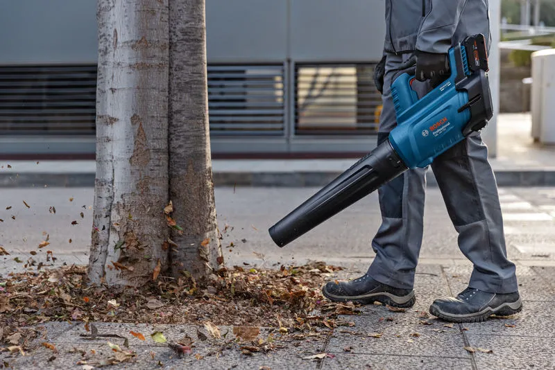 A person wearing safety equipment uses a cordless leaf blower to move leaves near a tree.