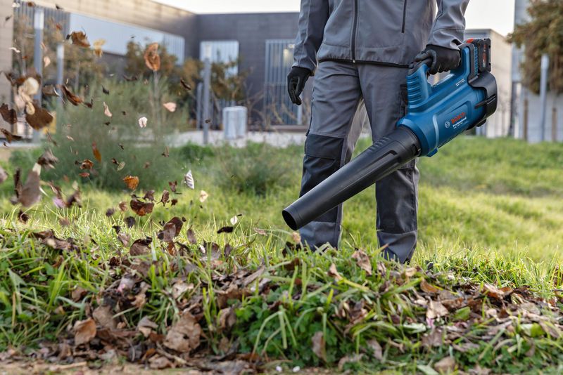 A person wearing safety equipment clears leaves with a cordless leaf blower outdoors.