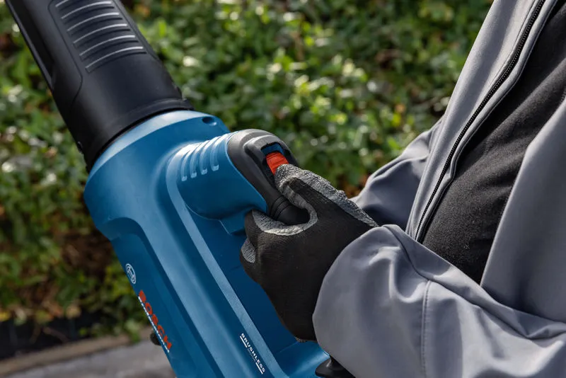 A person wearing safety equipment operates a cordless leaf blower outdoors.