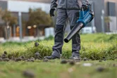 A person wearing safety equipment uses a cordless leaf blower on grass.
