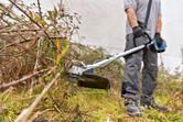 A person wearing safety equipment trims overgrown grass and brush with a cordless brushcutter.
