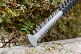 Hedge trimmer blade cutting green shrubbery along a garden edge.