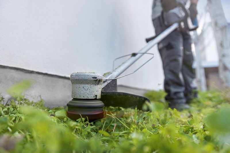 A person wearing safety equipment trims grass along a wall using a cordless grass trimmer.