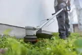 A person wearing safety equipment trims grass along a wall using a cordless grass trimmer.