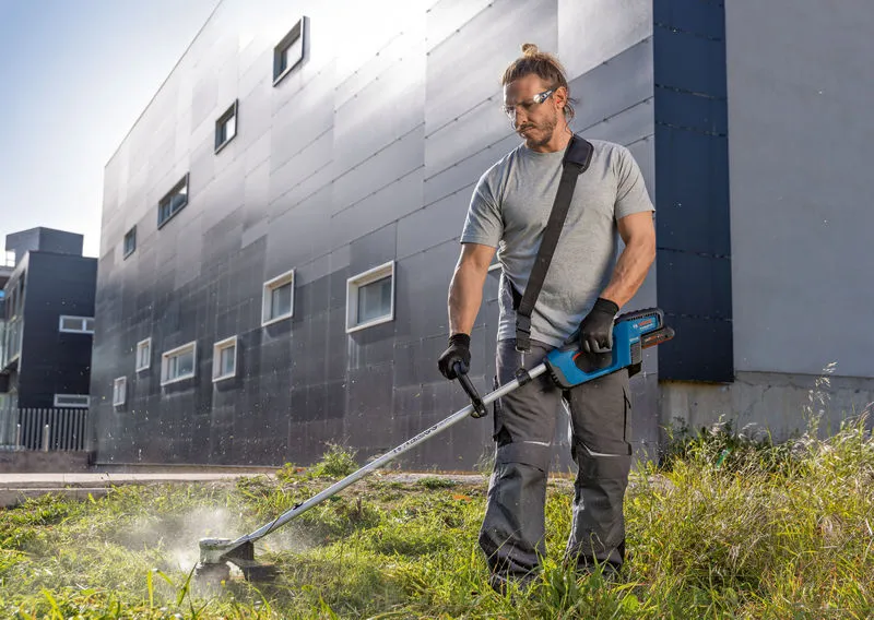 A person wearing safety equipment trims grass with a cordless grass trimmer outside a building.