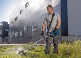 A person wearing safety equipment trims grass with a cordless grass trimmer outside a building.