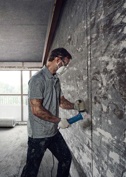 A person wearing safety equipment uses an angle grinder to cut into a concrete wall.
