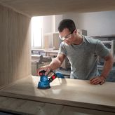 A person wearing safety equipment sands wood with a random orbit sander inside a workshop.