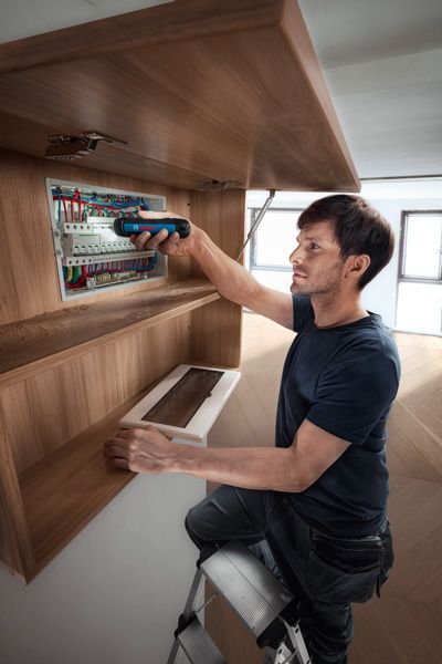 A person on a ladder uses a cordless screwdriver to work on an electrical panel.