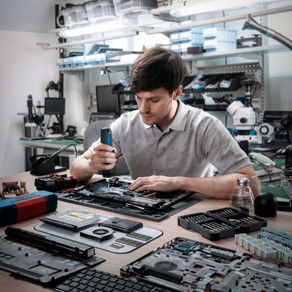 A person assembles electronics at a workbench using a cordless screwdriver.