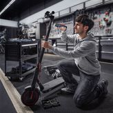 A person kneels and uses a cordless screwdriver to repair an electric scooter.