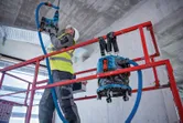 A person wearing safety equipment drills into a ceiling on a scaffold with a dust extractor nearby.