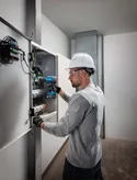 Person wearing safety equipment uses a cordless drill on an electrical panel.