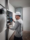 Person wearing safety equipment uses a cordless drill on an electrical panel.