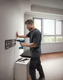 Person wearing safety equipment drills into a wall to install a metal mounting bracket.